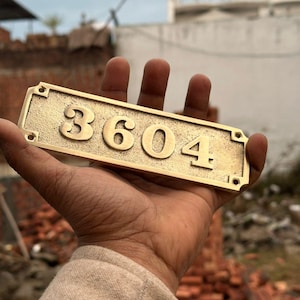 May include: A rectangular brass house number plaque featuring the raised digits 3604. The plaque has a textured, matte finish with a border and four pre-drilled holes at the corners for mounting, showcasing a metallic gold-toned appearance.