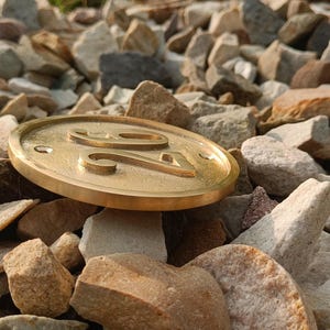 May include: A circular brass house number plaque featuring the embossed digits 24. The plaque has a smooth, metallic finish with a raised border and two small circular holes for mounting, positioned horizontally on a bed of various light-colored stones.