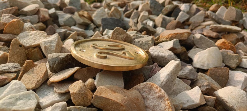 May include: A circular brass house number plaque featuring the embossed digits 24. The plaque has a smooth, metallic finish with a raised border and two small circular holes for mounting, positioned horizontally on a bed of various light-colored stones.