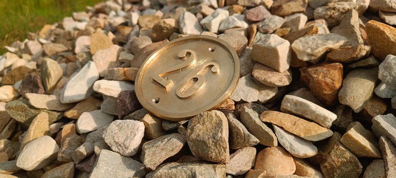 May include: A circular brass house number plaque featuring the embossed number 26 in a serif font. The plaque has a smooth, metallic finish with two small pre-drilled holes at the top and bottom for mounting purposes, resting on a bed of light-colored stones.