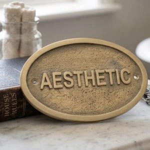 May include: An oval-shaped brass-toned metal plaque with a textured surface and raised lettering that reads AESTHETIC. The plaque features two small circular holes on the left and right sides for mounting purposes and has a weathered, antique finish.