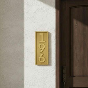 May include: A rectangular gold-colored house number plaque featuring the vertically aligned digits 1, 9, and 6 in a raised, sans-serif font. The plaque is mounted on a textured white wall next to a dark brown wooden door frame.