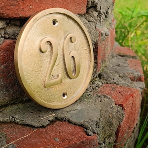 May include: A circular brass house number plaque featuring the embossed digits 26 in the center. The plaque has a smooth, metallic finish with two small pre-drilled screw holes positioned at the top and bottom for mounting purposes.