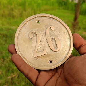 May include: A circular brass house number plaque featuring the embossed digits 26 in the center. The plaque has a smooth, metallic finish with two small pre-drilled holes at the top and bottom for mounting purposes, displaying a classic design.