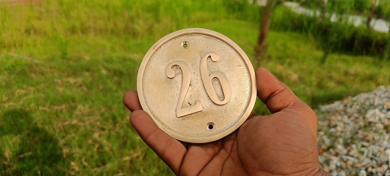 May include: A circular brass house number plaque featuring the embossed digits 26 in the center. The plaque has a smooth, metallic finish with two small pre-drilled holes at the top and bottom for mounting purposes, displaying a classic design.