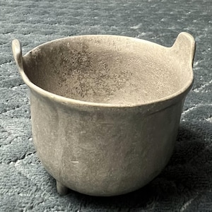 May include: A gray, ceramic cauldron with two handles and three small feet. The cauldron has a textured surface and a wide opening. The design is simple and rustic, suitable for decorative or functional use.