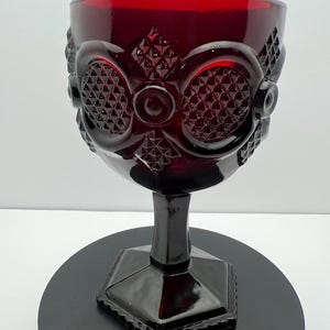 May include: A deep red glass goblet featuring a pressed pattern of circular and diamond-shaped motifs. The vessel has a thick stem and a hexagonal base with a beaded edge, showcasing a rich, translucent ruby color throughout the entire piece.