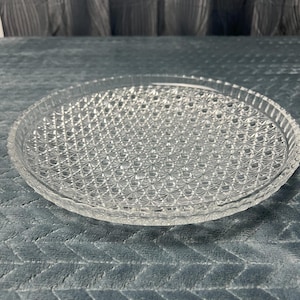May include: A clear, round glass serving plate with a diamond pattern across the surface. The plate has a slightly raised edge and is designed for serving food or displaying decorative items. The plate is on a blue-gray surface.
