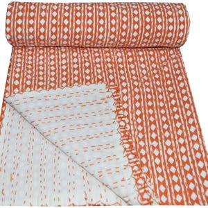 May include: Rolled-up orange and white patterned quilt. The quilt features a repeating pattern of vertical lines and diamond shapes in orange against a white background. The edges are scalloped, and the stitching is visible.