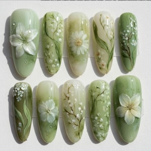 Puede incluir: Un conjunto de diez uñas artificiales con base verde claro y diseños florales. Cada uña presenta un diseño diferente pintado a mano, incluyendo flores blancas, margaritas y delicadas ramas con pequeñas flores blancas. Algunas uñas tienen una flor en 3D.