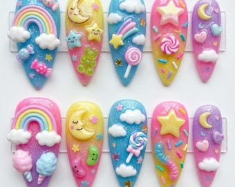 May include: A set of ten long, almond-shaped artificial nails. Each nail features a unique design with glitter, rainbows, clouds, stars, moons, candies, and other whimsical elements in pastel colors. The nails are arranged in two rows of five.