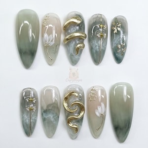 May include: A set of ten almond-shaped artificial nails. The nails feature a variety of designs, including gold snake and floral accents, and a marbled green and beige color scheme. The nails are arranged on a white surface.