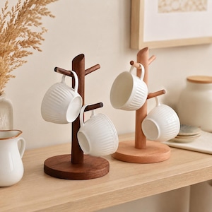 Può includere: Due supporti per tazze in legno con basi rotonde, uno con finitura marrone scuro e uno in legno naturale chiaro. Ogni supporto presenta diversi rami che sostengono tazze in ceramica bianca con una texture a coste verticali sulla superficie.
