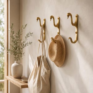 May include: Three gold-toned metal wall hooks shaped like swan necks, each featuring two hooks for hanging items. A beige canvas tote bag hangs from the first hook, and a tan straw hat hangs from the second hook, while the third hook remains empty.
