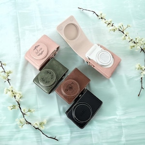 Può includere: Una collezione di custodie per fotocamere personalizzate in vari colori, tra cui rosa, verde, marrone e nero. Ogni custodia presenta un design circolare con testo o grafica personalizzati. Una custodia è aperta, rivelando una fotocamera bianca.