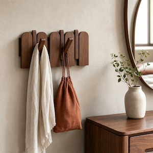 Puede incluir: Un perchero de pared de madera oscura con una serie de listones verticales arqueados. Dos ganchos de madera en ángulo sobresalen del soporte, sosteniendo una bufanda de lino beige claro y una bolsa de tela color óxido.
