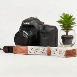 May include: A black Canon DSLR camera with a lens, paired with a cream-colored camera strap featuring embroidered floral patterns in blue and green. The strap has brown leather accents and the name "Julia" is printed on it.