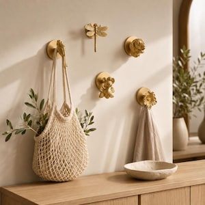 Puede incluir: Una colección de cinco ganchos de pared dorados con diseños esculpidos de un pájaro, una libélula, una cabeza de perro, un animal pequeño y una flor. Un gancho sostiene una bolsa de red beige y otro sostiene una toalla de tela beige en la pared.