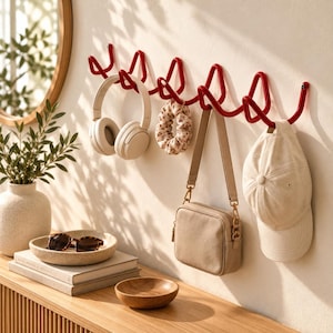 Puede incluir: Un perchero de pared de metal rojo con múltiples ganchos. Colgados de los ganchos hay un par de auriculares beige, una goma para el pelo de tela estampada, un bolso bandolera beige con correa y una gorra de béisbol de color beige.