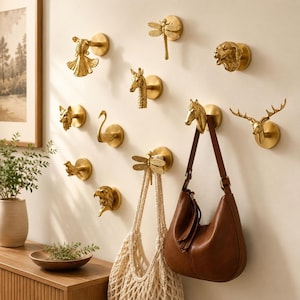 May include: A collection of gold-toned wall hooks shaped like various animals and insects, including a dragonfly, horse, deer, swan, eagle, fox, giraffe, dog, and fish. A brown leather shoulder bag and a cream-colored net bag hang from the hooks.