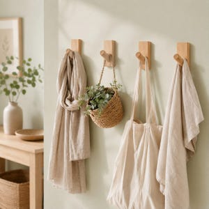 May include: Four light-colored wooden wall hooks mounted in a horizontal row. A beige linen scarf hangs from the first hook, a small woven basket with green foliage hangs from the second, a beige canvas tote bag hangs from the third, and a beige linen towel hangs from the fourth.