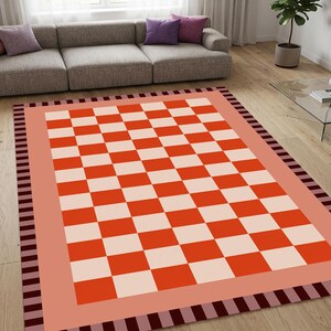 May include: A rectangular area rug featuring a central checkerboard pattern in vibrant orange and soft cream squares. The rug is framed by a solid peach-coloured border, which is enclosed by an outer edge of alternating dark brown and maroon stripes.