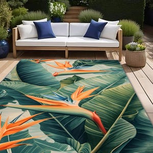 May include: A vibrant outdoor rug featuring a tropical bird of paradise floral design. The rug showcases large green leaves and orange, blue, and yellow flowers. The rug is placed on a wooden deck, with a white sofa and blue pillows in the background.