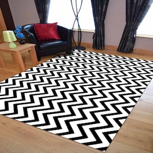 May include: A rectangular rug featuring a striking black and white zigzag design. The rug is laid on a wooden floor. A black armchair with a red cushion is in the background, alongside curtains.