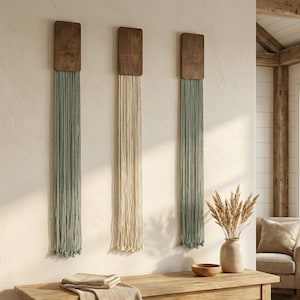 May include: Three wall hangings with wooden tops and long, fringed strands in sage green and cream. The decor is displayed on a light-colored wall, with a wooden console table below. A vase with dried wheat and a woven basket are also visible.