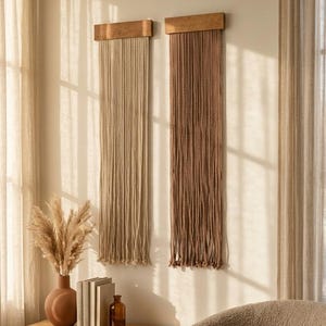 May include: Two vertical wall hangings featuring long, straight, hanging cords suspended from rectangular wooden bars. One wall hanging is light beige, and the other is a darker brown shade. Both pieces are mounted side-by-side on a plain wall.