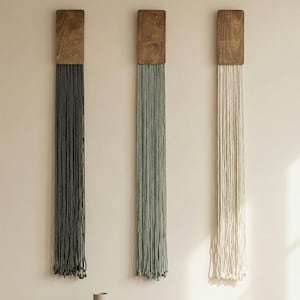 May include: Three wall hangings with wooden tops and hanging strands in gray, green, and off-white. Each piece features a rectangular wooden top with a series of knotted strands hanging below, creating a textured, decorative effect.