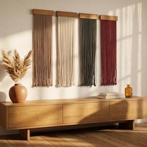 May include: Four vertical wall hangings featuring long, hanging cords in tan, cream, dark green, and deep red, each attached to a rectangular wooden top bar. These textile pieces are mounted side-by-side on a wall above a long wooden console table.