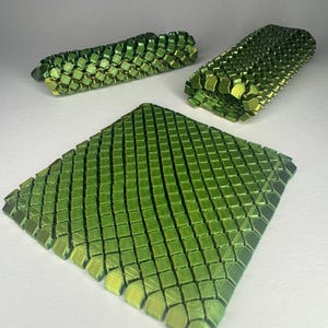Könnte beinhalten: Drei 3D-gedruckte Objekte mit schuppenartiger Textur. Die Objekte sind in Grün- und Gelbtönen gehalten und weisen ein quadratisches Kachelmuster auf. Ein Objekt ist ein flaches Quadrat, die anderen beiden sind gebogen.