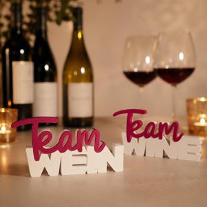 Könnte beinhalten: Zwei dekorative Tischaufsteller mit dem Text Team WEIN und Team WINE in einem geschichteten Design. Das Wort Team ist in einer dunkelmagentafarbenen Schreibschrift gehalten, während die Wörter WEIN und WINE in weißer Blockschrift sind.