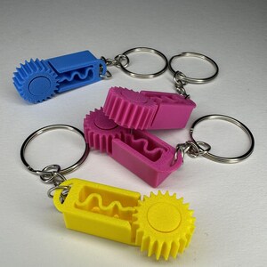 Può includere: Tre portachiavi colorati, blu, rosa e giallo. Ogni portachiavi presenta un design a forma di ingranaggio e un interno ondulato, collegato a un portachiavi argentato. I portachiavi sono realizzati in un materiale solido e sono su sfondo bianco.