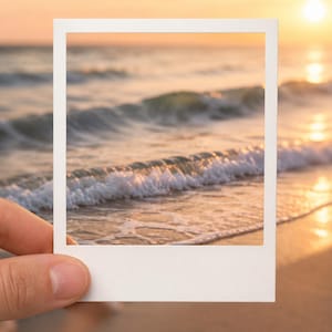 Op de afbeelding: Een witte vierkante fotolijst die voor een strandscène bij zonsondergang wordt gehouden. De lijst toont oceaangolven die op het zandstrand rollen met gouden zonlicht dat op het water reflecteert. De tekst digital download is zichtbaar.