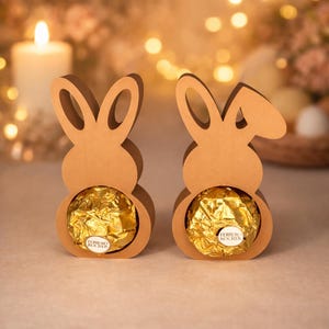 Puede incluir: Dos soportes de cartón color canela con forma de conejo, cada uno con un chocolate Ferrero Rocher redondo envuelto en papel dorado. El texto digital download es visible en la esquina superior derecha. Los conejos tienen orejas recortadas.
