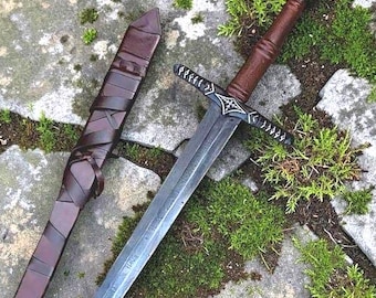 Puede incluir: Una espada de estilo medieval con una hoja plateada y un mango marrón, junto a una vaina de cuero marrón. La espada tiene una empuñadura en forma de cruz y está sobre una superficie de piedra con musgo verde.