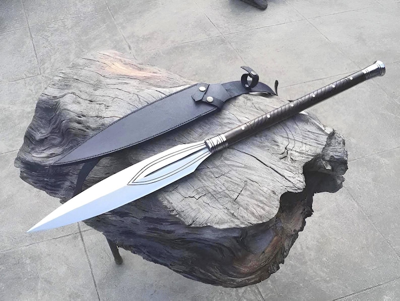 Puede incluir: Una lanza con una hoja plateada y un mango marr&oacute;n oscuro, apoyada sobre una superficie de madera envejecida. La lanza va acompa&ntilde;ada de una funda de cuero negro. La hoja de la lanza tiene un dise&ntilde;o decorativo.