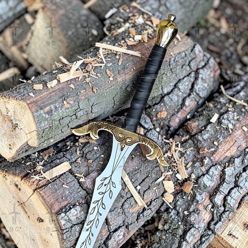 Puede incluir: Una espada decorativa con empu&ntilde;adura y guarda doradas, un mango envuelto en negro y una hoja plateada. La hoja presenta un dise&ntilde;o de hojas. La espada descansa sobre una pila de troncos.