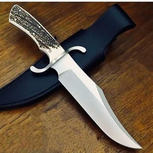 May include: A large hunting knife with a polished steel blade and a handle made of bone-like material with dark brown accents. The knife rests in a black leather sheath on a wooden surface.