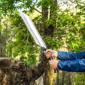 Könnte beinhalten: Eine große, silberfarbene Klinge mit einem dunkelbraunen Holzgriff. Die Klinge wird von zwei Händen gehalten, mit einem Hintergrund aus grünem Laub und Bäumen. Die Klinge scheint ein Schwert oder ein großes Messer zu sein.