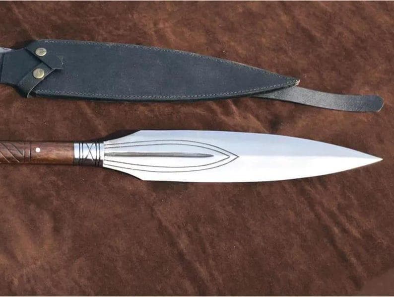 Puede incluir: Una lanza con una hoja de acero pulido y un mango de madera. El mango tiene un dise&ntilde;o tallado y est&aacute; conectado a la hoja con un metal de color plateado. Se incluye una funda de cuero negro.