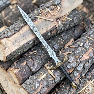 Puede incluir: Una espada plateada con un dise&ntilde;o de hojas grabado en la hoja, una guarda dorada y un mango negro envuelto. La espada se exhibe sobre un lecho de troncos de madera.