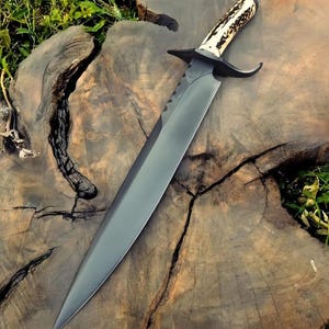 May include: A large, black hunting knife with a polished blade and a handle made of bone and dark accents. The knife features a guard and a serrated section near the handle. The knife is displayed on a weathered wooden surface.
