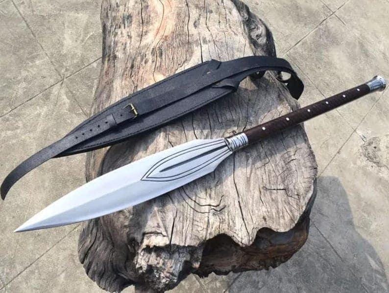 Puede incluir: Una lanza con una hoja plateada y un mango marr&oacute;n oscuro con puntos blancos. La lanza est&aacute; sobre una superficie de madera desgastada, con una funda y correa de cuero negro.