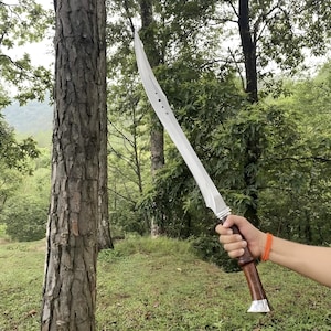 Könnte beinhalten: Ein großes, gebogenes Schwert mit einer silbernen Klinge und einem dunklen Holzgriff. Der Griff hat einen silbernen Sockel und einen dunkelbraunen Griff. Das Schwert wird vor einem Baum und grünem Laub gehalten.