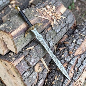 Puede incluir: Una espada decorativa con una hoja plateada y una empu&ntilde;adura dorada, apoyada sobre una pila de troncos de madera. La hoja presenta un dise&ntilde;o de hojas grabado. El mango est&aacute; envuelto en material negro.