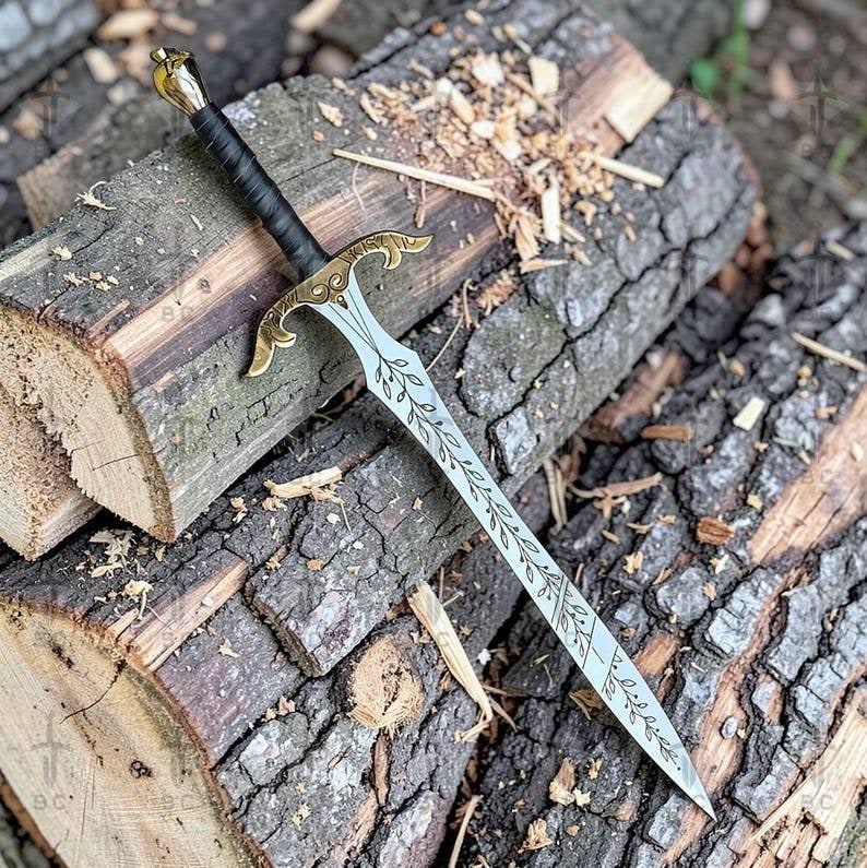 Puede incluir: Una espada decorativa con una hoja plateada y una empu&ntilde;adura dorada, apoyada sobre una pila de troncos de madera. La hoja presenta un dise&ntilde;o de hojas grabado. El mango est&aacute; envuelto en material negro.