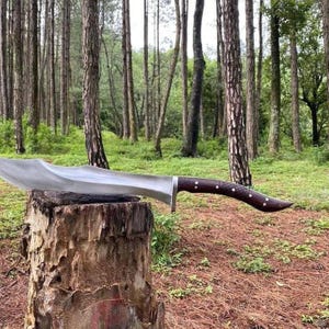 May include: A large, curved blade knife with a polished steel finish and a dark brown wooden handle. The knife is resting on a tree stump in a forest setting. The handle has decorative white dots.